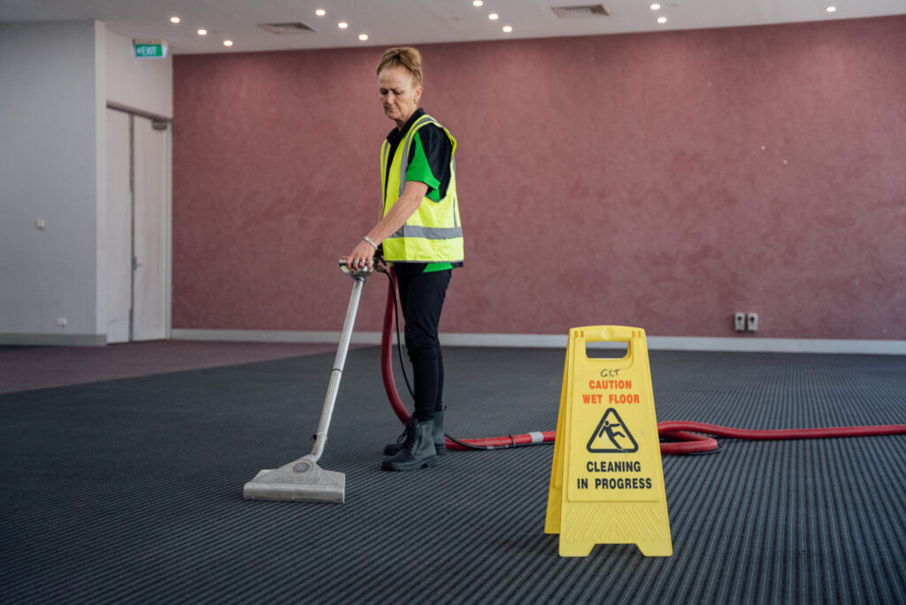 A Green Clean Team member cleaning carpets in front of a yellow 'cleaning in progress' sign. The person is performing an eco-friendly carpet cleaning job in Bendigo, Central Victoria.